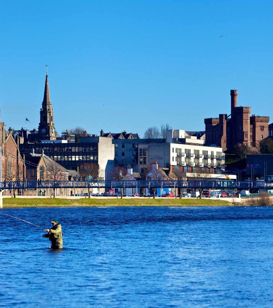 A man fishing in River Ness with the city of Inverness in the background