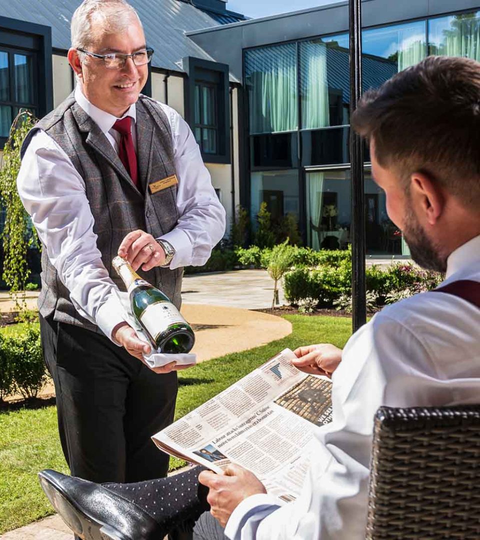 Serving a bottle of champagne in courtyard to guest reading newspaper