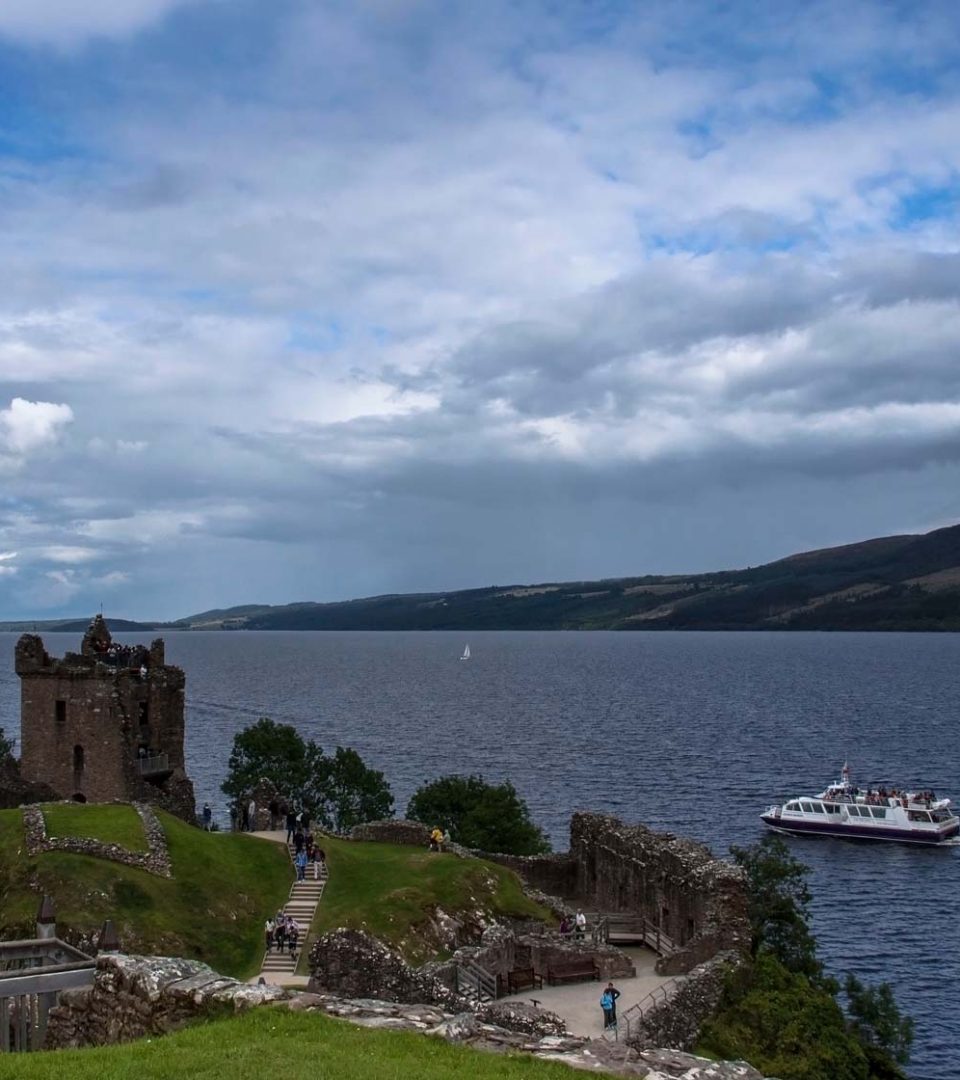 Urquhart Castle with Loch Ness in the background and a Jacobite boat tour passing by.