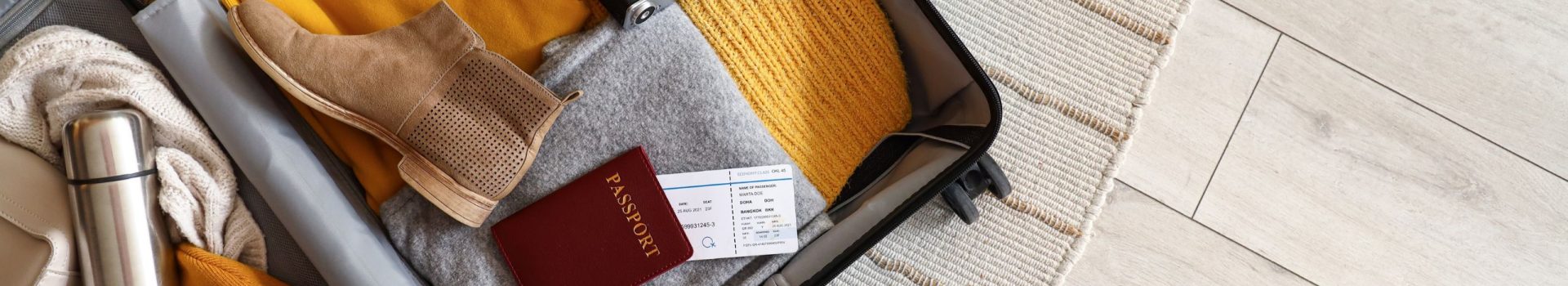 Open suitcase with travelling accessories and winter clothes on carpet, top view.