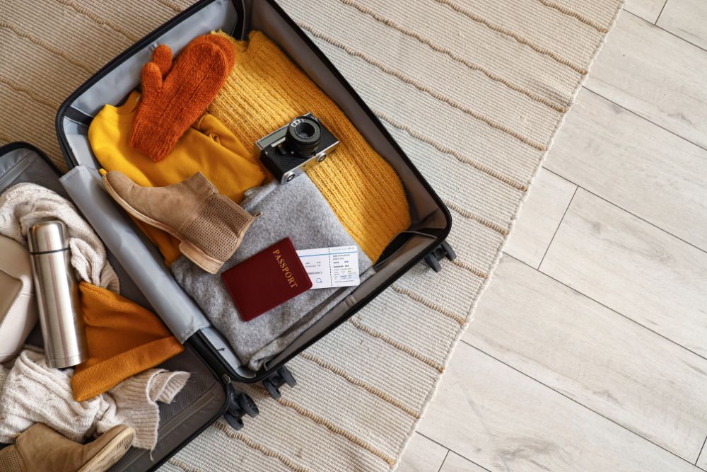 Open suitcase with travelling accessories and winter clothes on carpet, top view.