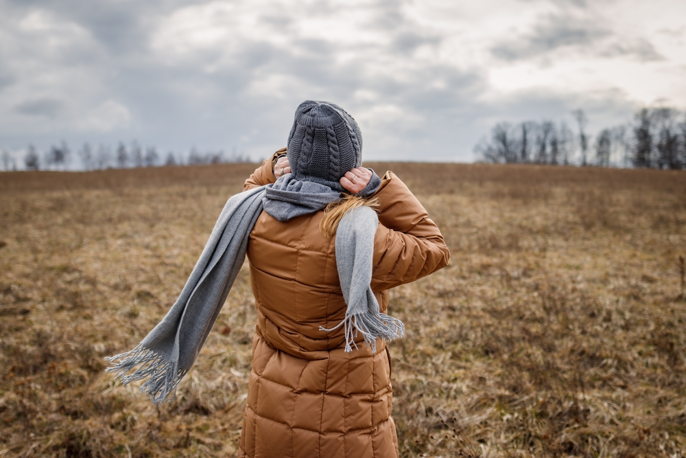 Wind and cold weather. Woman wearing coat, scarf and knit hat outdoors. Female person walks in nature.